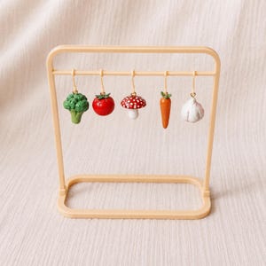Könnte beinhalten: Ein beigefarbener Kunststoffständer präsentiert fünf Miniatur-Anhänger in Form von Gemüse und Pilzen. Die Anhänger bestehen aus einem grünen Brokkoli, einer roten Tomate, einem rot-weiß gepunkteten Pilz, einer orangefarbenen Karotte und einer weißen Knoblauchknolle.