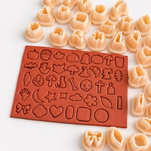 Può includere: Uno stampo in silicone rettangolare color terracotta con una griglia di varie piccole forme, tra cui zucche, teschi, stelle, cuori e fiocchi, accompagnato da una collezione di formine per biscotti in plastica beige abbinate.