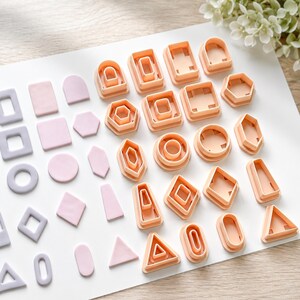 May include: A collection of peach-coloured plastic geometric clay cutters arranged alongside various light purple and pink clay shapes, including squares, circles, triangles, hexagons, and arches, displayed on a white surface.