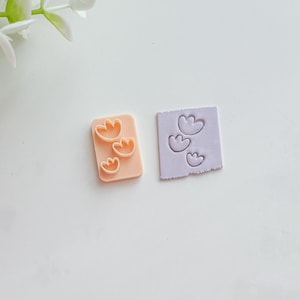 Può includere: Due piccoli tagliapasta rettangolari in argilla. Uno è color pesca con tre forme di fiori, e l'altro è lavanda con tre impronte di fiori. I tagliapasta sono per l'artigianato o la cottura.