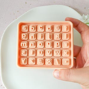 Op de afbeelding: Een perzikkleurige siliconen alfabetmal met 26 letterstempels, cijfers en symbolen. De mal wordt door een hand vastgehouden en ligt op een wit bord. De letters zijn in een schreefloze lettertype.