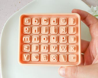 May include: A peach-coloured silicone alphabet mould with 26 letter stamps, numbers, and symbols. The mould is held by a hand and sits on a white plate. The letters are in a sans-serif font.