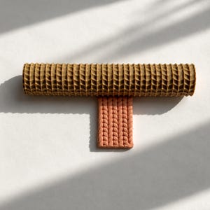 Puede incluir: Un rodillo cilíndrico con una superficie marrón de textura en cuadrícula colocado horizontalmente sobre un bloque rectangular de color melocotón con un patrón vertical de tejido trenzado. Ambos objetos tienen un aspecto mate y arcilloso.