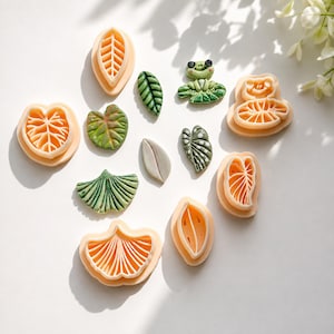 Può includere: Una collezione di formine in plastica color pesca a forma di foglie e rane, insieme a diversi pezzi di argilla verde e bianca finiti e una piccola statuetta di rana verde, disposti su una superficie bianca.