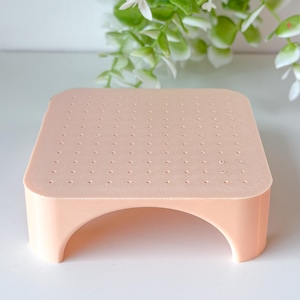 May include: A peach-colored, square-shaped cake pop stand with a perforated top and arched supports. The stand is designed to hold multiple cake pops, with evenly spaced holes for inserting the sticks. The stand is made of a solid, durable material.