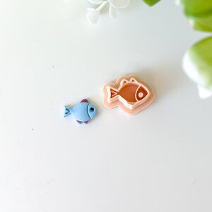Può includere: Una statuetta a forma di pesce azzurro con dettagli rossi e viola accanto a un tagliabiscotti a forma di pesce color pesca. Gli oggetti sono su una superficie bianca, con accenti floreali verdi e bianchi sullo sfondo.