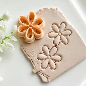 Op de afbeelding: Een perzikkleurige plastic bloemvormige uitsteker op een plat stuk lichtbeige klei. De uitsteker heeft zes afgeronde bloemblaadjes en een centrale ronde kern. Twee identieke bloemafdrukken zijn duidelijk zichtbaar in de klei.