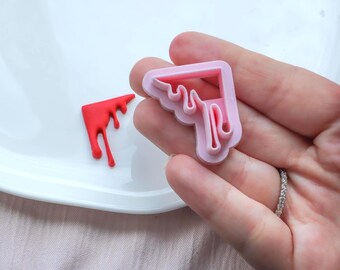 May include: A pink plastic cookie cutter in the shape of a dripping corner, held in a hand. A small red, corner-shaped biscuit with a dripping design sits on a white plate.