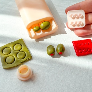 Op de afbeelding: Een verzameling miniatuurvoedsel, waaronder groene olijven met rode uiteinden, een rechthoekig blok met cirkelvormige inkepingen en een groen vierkant met cirkelvormige afdrukken. De items zijn gerangschikt op een wit oppervlak.