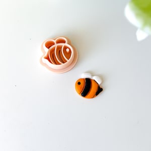 May include: A light pink bee-shaped cookie cutter next to a small, decorated bee. The bee is orange with black stripes and white wings. The cookie cutter is made of plastic and has a detailed bee design.