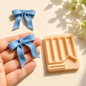 May include: Two light blue bows with a textured, ribbed surface and a beige rectangular plastic mold featuring cutouts for creating bow shapes. The bows have a central knot and two hanging tails, while the mold has distinct sections.
