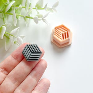 Könnte beinhalten: Ein grauer geometrischer Polymer Clay-Ohrring mit gewebtem Design wird in einer Hand gehalten. Ein hellpfirsichfarbener Polymer Clay-Ohrringausstecher mit ähnlichem Design befindet sich in der Nähe. Der Hintergrund ist weiß mit grün-weißen floralen Akzenten.