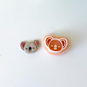 Peut inclure: Un visage de koala gris et un emporte-pièce en forme de koala. Le visage du koala a des oreilles roses et une expression souriante. L'emporte-pièce est rose clair et a la forme d'un visage de koala avec une expression souriante.