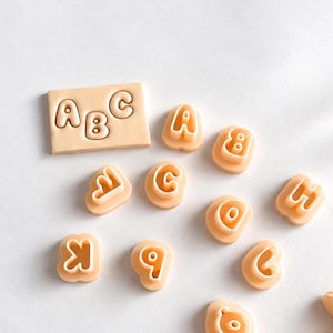 Könnte beinhalten: Ein Set aus hellpfirsichfarbenen Kunststoff-Ausstechformen für das Alphabet. Die Sammlung enthält eine rechteckige Platte mit den geprägten Buchstaben A, B und C sowie einzelne Ausstechformen für die Buchstaben A, B, C, D, H, K, Q und Z.