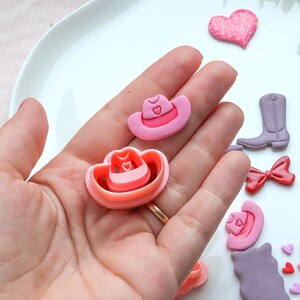 Könnte beinhalten: Eine Sammlung von rosa und roten Cowboy-Themen-Ausstechern und Dekorationen. Das Set enthält einen Cowboyhut, eine herzförmige Glitzerdekoration, einen Cowboy-Stiefel und eine rote Schleife. Der Cowboyhut-Ausstecher hat eine Herzform.