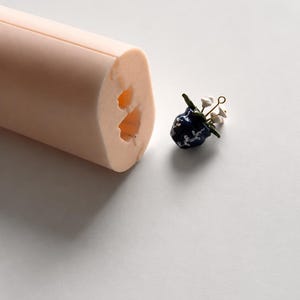 May include: A pale peach-coloured silicone mould with a hollowed-out interior shape and a small, dark blue ceramic charm featuring white floral details, green leaves, and a gold-toned metal loop at the top for attachment to jewellery or accessories.