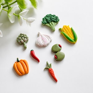 May include: A collection of small, colourful vegetable-shaped charms including a green broccoli floret, a yellow corn cob, a white garlic bulb, a green artichoke, two green olives with red centres, a red chilli pepper, an orange pumpkin, and a carrot.