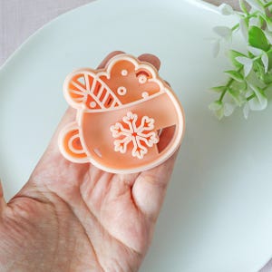 Könnte beinhalten: Hellorangefarbener Ausstecher aus Kunststoff in Form einer Tasse heißer Schokolade mit Schneeflocken-Design. Der Ausstecher hat einen Griff und wird in einer Hand gehalten. Die Tasse ist mit Schlagsahne und einem Zuckerstange verziert.