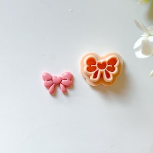 Op de afbeelding: Twee roze strikvormige koekjessnijders. Eén is een effen strik en de andere is een snijder met een strikdesign. De snijder heeft een hartvormig midden. De items liggen op een wit oppervlak.