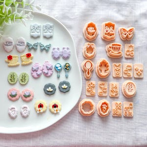 Op de afbeelding: Een verzameling decoratieve ambachtelijke mallen en versieringen in verschillende vormen en kleuren. De collectie bevat bloemen, vlinders en andere op de natuur geïnspireerde ontwerpen, samen met een verscheidenheid aan koekjessnijders.