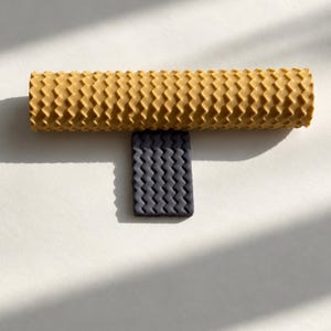 Puede incluir: Una herramienta de rodillo cilíndrica de color amarillo mostaza con un patrón de textura en zigzag repetitivo, colocada horizontalmente sobre un pequeño bloque rectangular de color gris oscuro con el mismo patrón en zigzag.