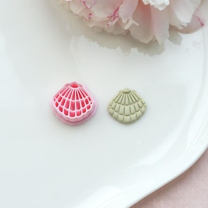 Può includere: Due formine per biscotti a forma di conchiglia. Una è rosa con un motivo a griglia, l'altra è verde chiaro con un design a coste. Le formine sono su un piatto bianco, con un fiore rosa sullo sfondo.