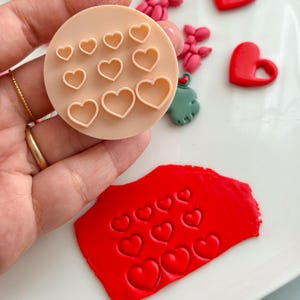 Op de afbeelding: Een lichtbeige hartvormige koekjessnijder met verschillende maten hartuitsparingen. Een rood stuk klei met hartindrukken ligt naast de snijder. Andere hartvormige decoraties zijn ook zichtbaar.