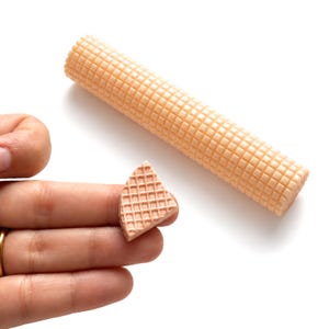 May include: A cylindrical, light beige wafer roll featuring a textured grid pattern on its surface, accompanied by a small, triangular-shaped piece of the same wafer material, both displaying a consistent waffle-like embossed design.