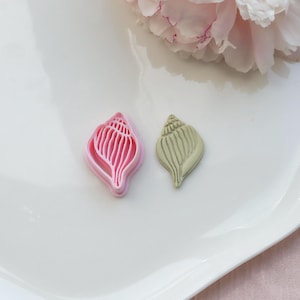 Könnte beinhalten: Ein rosa muschelförmiger Ausstecher neben einem muschelförmigen Keks. Der Ausstecher besteht aus rosa Kunststoff und hat ein detailliertes Design. Der Keks ist hellgrün und hat eine strukturierte Oberfläche.