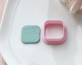 Può includere: Un tagliabiscotti quadrato rosa chiaro accanto a un quadrato verde chiaro strutturato. Il tagliabiscotti è aperto, mostrando l'interno. Gli oggetti sono su un piatto bianco.
