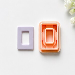 Può includere: Un tagliapasta rettangolare color viola chiaro con superficie liscia e uno stampo rettangolare color pesca abbinato, dotato di un telaio interno rialzato e nervature di supporto strutturale, progettato per modellare materiali in forme rettangolari.