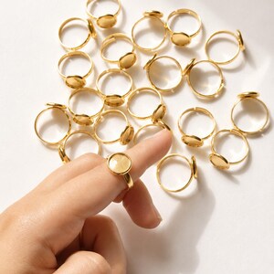 May include: A collection of gold-toned metal ring blanks, each featuring a circular flat bezel setting for crafting jewelry. The rings have thin, adjustable bands and a polished finish. One ring is shown positioned on a finger for scale.