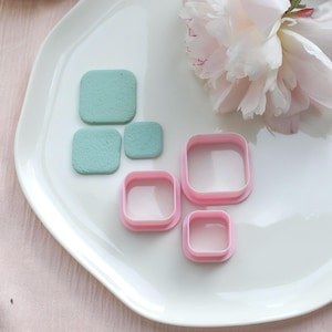 Può includere: Un piatto bianco contiene tre oggetti quadrati azzurri e tre formine per biscotti quadrate rosa. Sullo sfondo, una grande peonia rosa chiaro. Gli oggetti sono disposti su una superficie bianca leggermente strutturata.