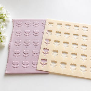 Op de afbeelding: Een rechthoekig mauve vel materiaal met een herhalend reliëfpatroon van kleine bloemen, gedeeltelijk bedekt door een crèmekleurig sjabloon met een bijpassend raster van uitgesneden bloemvormen gerangschikt in nette rijen en kolommen.