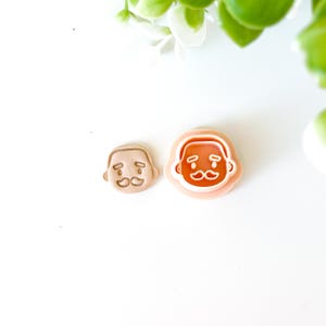 Peut inclure: Deux emporte-pièces en forme de visage de Père Noël. L'un est un biscuit de couleur beige, et l'autre est un découpoir en plastique rose. Les visages du Père Noël ont une moustache et une expression souriante. Le fond est blanc avec un feuillage vert et blanc.