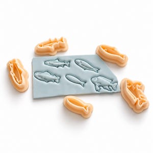 Puede incluir: Un molde de silicona rectangular de color azul claro con cinco hendiduras distintas en forma de pez, rodeado por cinco cortadores de galletas de plástico de color melocotón que coinciden con las formas de las impresiones del molde.