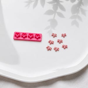 Könnte beinhalten: Eine rechteckige, rosafarbene Silikonform mit vier identischen blattförmigen Vertiefungen, begleitet von sechs kleinen, altrosa blattförmigen Verzierungen, die aus der Form gegossen wurden und detaillierte Blattadern sowie spitze Ränder aufweisen.