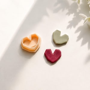 May include: A peach-coloured geometric heart-shaped cutter alongside two finished heart-shaped pieces, one in a muted sage green and the other in a deep, speckled burgundy red, all featuring sharp, angular edges and a flat, symmetrical design.