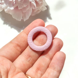 Puede incluir: Una cuenta de madera circular de color rosa claro con una superficie texturizada. La cuenta se sostiene en la mano de una persona, con una flor rosa borrosa en el fondo. La cuenta tiene aproximadamente 2,5 cm de diámetro.