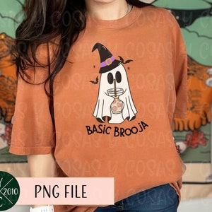 Könnte beinhalten: Ein orangefarbenes T-Shirt mit einem weißen Geist, der einen Hexenhut trägt und ein Getränk hält. Der Text "Basic Brooja" ist unter dem Geist gedruckt.