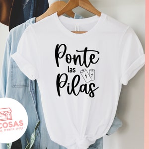Op de afbeelding: Wit T-shirt met zwarte tekst die "Ponte las Pilas" zegt met twee batterijpictogrammen. De tekst is in een cursief schrift. Het T-shirt bevindt zich op een blauwe denim achtergrond.