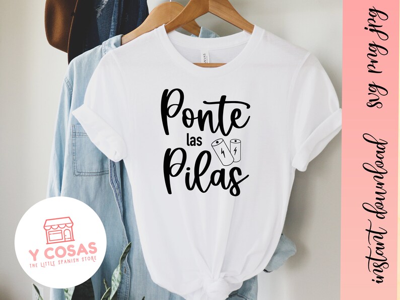 Op de afbeelding: Wit T-shirt met zwarte tekst die "Ponte las Pilas" zegt met twee batterijpictogrammen. De tekst is in een cursief schrift. Het T-shirt bevindt zich op een blauwe denim achtergrond.
