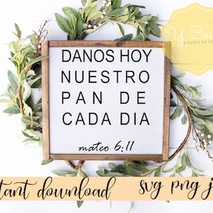 May include: A white sign with a wooden frame, featuring the text "DANOS HOY NUESTRO PAN DE CADA DIA mateo 6:11" in black lettering. The sign is surrounded by a green leafy garland.