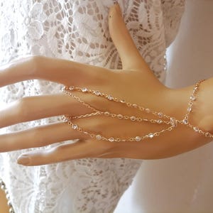 May include: A delicate gold hand chain with clear crystal accents. The jewelry piece features multiple strands that connect to the fingers and wrist, creating an elegant and sophisticated look. The hand chain is displayed on a mannequin hand.