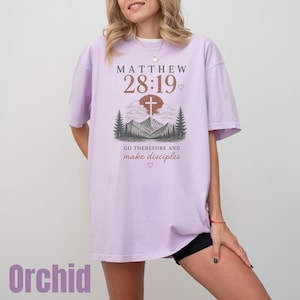 May include: An orchid-colored t-shirt featuring a graphic print with the text Matthew 28:19, a cross, mountains, and pine trees. Below the illustration, the text reads Go therefore and make disciples, accompanied by small heart icons.