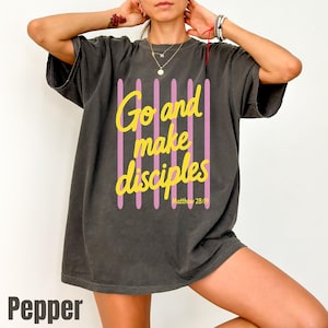 May include: A charcoal gray short-sleeve t-shirt featuring a graphic design with vertical light pink stripes and the yellow text "Go and make disciples" followed by "Matthew 28:19" in a smaller yellow font centered on the front of the garment.