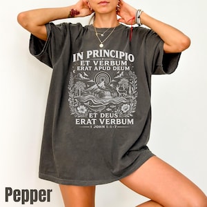 Peut inclure: Un t-shirt gris anthracite à manches courtes avec une impression graphique blanche représentant un paysage avec une montagne, une rivière, un cerf et une baleine. Le texte indique IN PRINCIPIO ET VERBUM ERAT APUD DEUM ET DEUS ERAT VERBUM 1 JOHN 1:1-7.