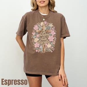 May include: A brown short-sleeve t-shirt featuring a graphic of a wooden cross surrounded by colorful pink, yellow, and blue flowers. The text "Go therefore and make disciples" is printed in a cursive font over the center of the cross design.