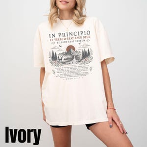 Peut inclure: Un t-shirt à manches courtes ivoire avec une impression graphique comprenant le texte latin IN PRINCIPIO ET VERBUM ERAT APUD DEUM ET DEUS ERAT VERBUM, un paysage de montagne avec un cerf, des pins, un soleil et le texte JOHN 1:1-7 en bas.