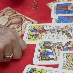 May include: A selection of Tarot cards with vibrant illustrations are arranged on a red surface. The cards display images and French text. A person wearing a gold ring is holding the cards.